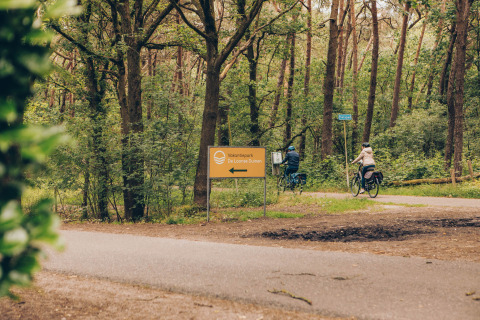 Two cyclists ride through a forest near Dune Lodge at Duinhoeve, Netherlands, passing a holiday park sign.