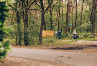 Two cyclists ride through a forest near Dune Lodge at Duinhoeve, Netherlands, passing a holiday park sign.
