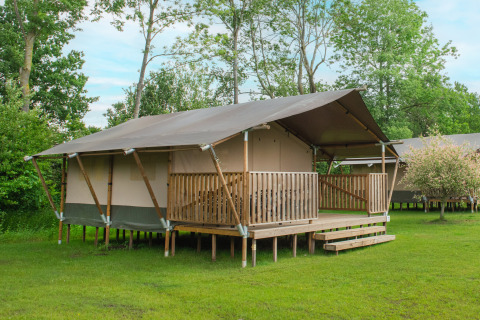 Safarizelt mit Holzveranda auf grüner Wiese, umgeben von Bäumen, perfekt für luxuriöses Camping.