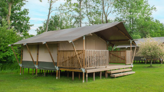 Safarizelt mit Holzveranda auf grüner Wiese, umgeben von Bäumen, perfekt für luxuriöses Camping.