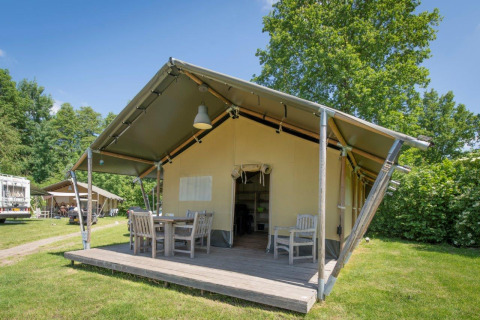 Außenaufnahme eines Safarizeltes mit Holzmöbeln auf der Veranda, umgeben von grünem Gras und Bäumen.
