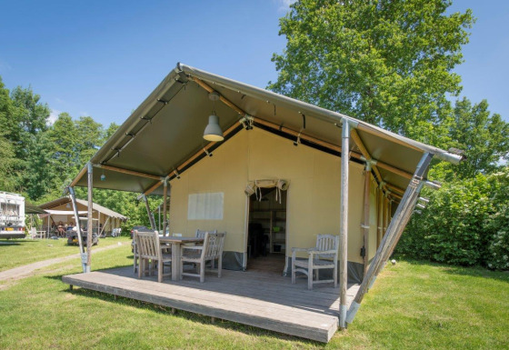 Außenaufnahme eines Safarizeltes mit Holzmöbeln auf der Veranda, umgeben von grünem Gras und Bäumen.