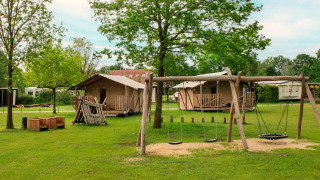 Safaritent camping met speeltoestellen en houten huisjes omringd door groene bomen en gras.