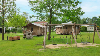 Safari-Zeltplatz mit Spielplatz und Holzhütten unter grünen Bäumen, ideal für Familienurlaub.
