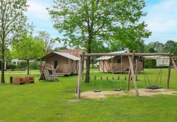 Safari-Zeltplatz mit Spielplatz und Holzhütten unter grünen Bäumen, ideal für Familienurlaub.