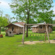 Espace camping avec tente safari, aire de jeux et cabanes en bois sous de grands arbres verts.
