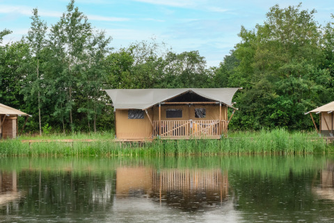 Safari tent by the lake with sanitary facilities, surrounded by trees and reflected in calm water.