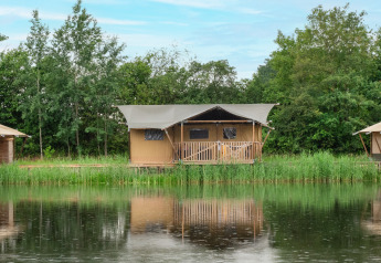 Safari telt ved søen med sanitære faciliteter, omgivet af træer og refleksion i det rolige vand.