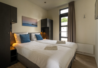 Moderne tweepersoonskamer in Villa Maritiem lodge met witte lakens, blauwe kussens en ramen met uitzicht.
