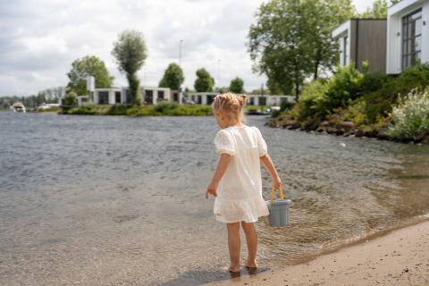 Bambina con abito bianco e secchiello in riva all’acqua davanti alle ville di MarinaPark Bad Nederrijn.