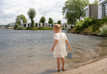 Bambina con abito bianco e secchiello in riva all’acqua davanti alle ville di MarinaPark Bad Nederrijn.