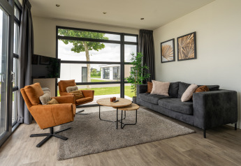 Moderne woonkamer met grijze zetel, oranje stoelen en grote ramen in Villa Maritiem, Nederland.