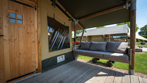 Area salotto esterna con divano e porta in legno alla tenda safari 'Compact' presso il Camping de Molenhoek, Olanda.