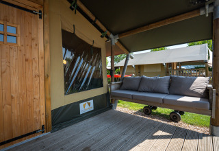 Buiten zitruimte met sofa en houten deur aan safaritent ‘Compact’ op Camping de Molenhoek, Nederland.