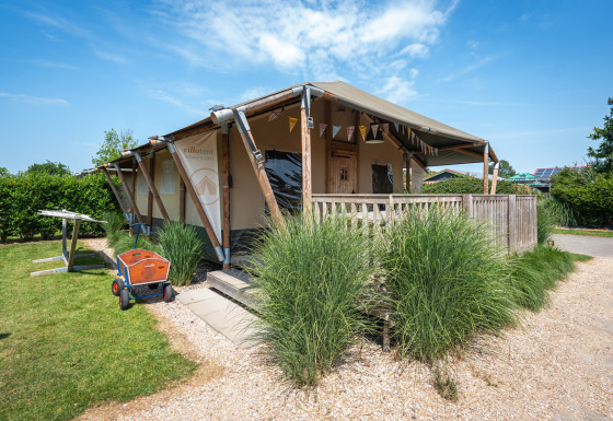 Tenda safari chiamata Villatent Cottage con veranda in legno, circondata dal verde sotto cielo blu.