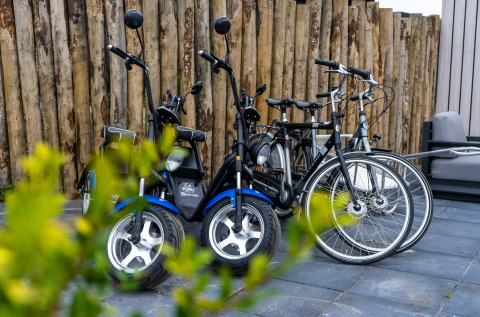 Biciclette e scooter elettrici parcheggiati sul patio di un lodge, con una recinzione in legno e piante davanti.