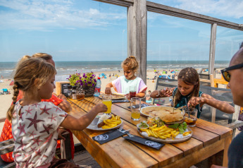 Niños almorzando en una mesa de madera en un restaurante junto a la playa con vista al mar en un día soleado.
