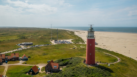 Aerial view of a red lighthouse by a sandy beach, green fields, small houses, and distant parked cars.