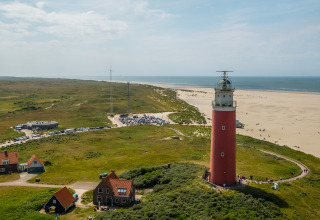 Aerial view of a red lighthouse by a sandy beach, green fields, small houses, and distant parked cars.