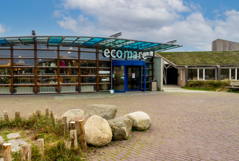Entrance of Ecomare lodge showing a modern glass facade, green roof, and large stones beneath blue sky.