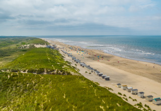A wide sandy beach with grassy dunes and beach cabins near a lodge, ocean waves and sky in the distance.