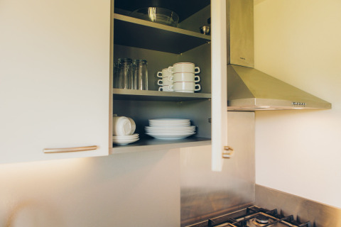 Open kitchen cabinet in a tiny house at Valleichalet with stacked cups, glasses and plates near stove.