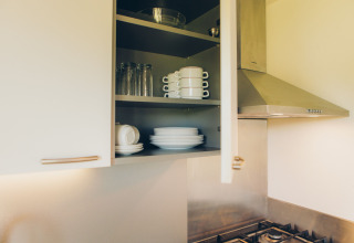 Open keukenkastje in een tiny house bij Valleichalet met stapels servies, glazen en kopjes naast het fornuis.
