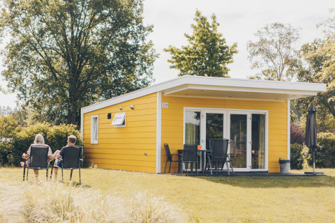 Two people relaxing outside a yellow Valleichalet tiny house at Holiday park Ackersate in the Netherlands.