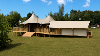 Safari tent Glamfield Lodge Deluxe met hot tub op een grasveld, omgeven door bomen en blauwe lucht.