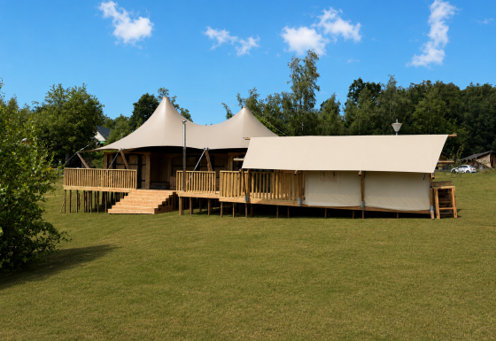 Safari tent Glamfield Lodge Deluxe met hot tub op een grasveld, omgeven door bomen en blauwe lucht.