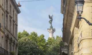 Vista de calle cerca de Audenge, Francia, con una estatua sobre una columna entre edificios históricos.