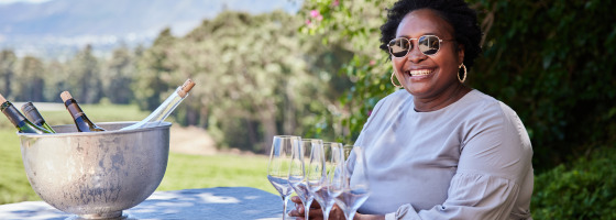 Vrouw met zonnebril lacht aan een tafel met wijn en glazen, buiten bij een glamping vakantiepark.