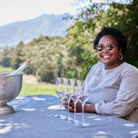 Mujer sonriente con gafas de sol junto a vino y copas en un parque de vacaciones glamping con paisaje.