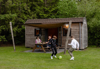 Gezin bij houten lodge op Camping de Noetselerberg in Nederland, kinderen spelen voetbal op het grasveld.