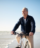 Hombre mayor monta en bicicleta por la playa en un parque vacacional con opciones de glamping al sol.