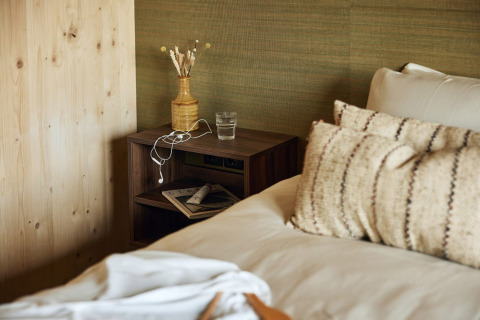 Habitación de lodge acogedora con cama, cojines y mesita con vaso de agua, jarrón y auriculares.