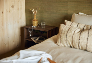 Habitación de lodge acogedora con cama, cojines y mesita con vaso de agua, jarrón y auriculares.