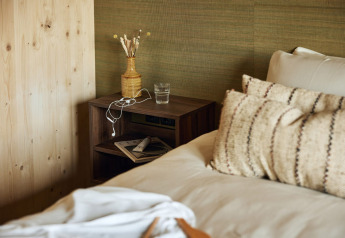 A cozy lodge room with a bed and cushions beside a wooden nightstand with water, vase, and earbuds.