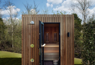 Sauna exterior de madera en un lodge, puerta abierta y cielo azul con nubes, entorno natural y verde.