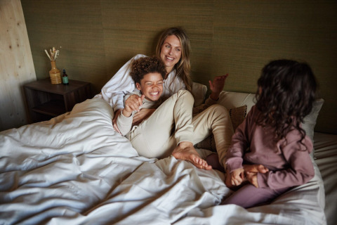 Una familia se relaja sobre una cama en una cabaña, sonriente y disfrutando juntos de un momento acogedor.