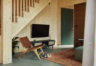 Salon moderne à la Unbrick One lodge avec chaise en bois, TV sous l'escalier et porte verte aux Pays-Bas.