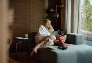 Donna e bambino rilassati su un divano in un'elegante lodge con grandi finestre a Brinckerduyn.