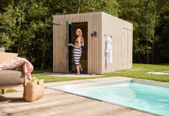 Mujer con toalla entrando a una sauna junto a la piscina en Brinckerduyn, Países Bajos, rodeada de árboles.