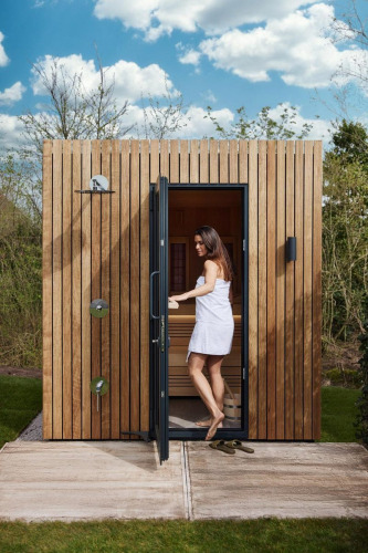Mujer con toalla sale de una moderna sauna de madera en glamping, rodeada de naturaleza y cielo azul.