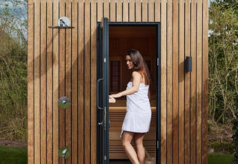 Mujer con toalla sale de una moderna sauna de madera en glamping, rodeada de naturaleza y cielo azul.