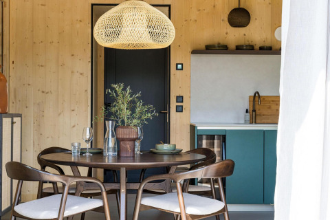 Comedor moderno en una cabaña, con paredes de madera, mesa redonda, sillas, lámpara colgante y cocineta.