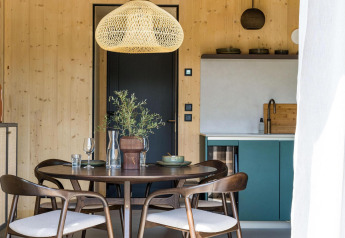 Comedor moderno en una cabaña, con paredes de madera, mesa redonda, sillas, lámpara colgante y cocineta.