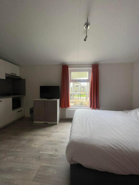 Lichte kamer in Comfort Lodge met tweepersoonsbed, televisie, raam met rode gordijnen en kitchenette.