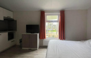 Lichte kamer in Comfort Lodge met tweepersoonsbed, televisie, raam met rode gordijnen en kitchenette.