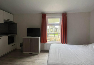 Lichte kamer in Comfort Lodge met tweepersoonsbed, televisie, raam met rode gordijnen en kitchenette.